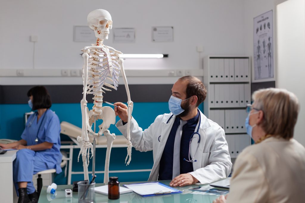 specialist explaining human skeleton retired woman checkup visit talking about bones injury osteopathy appointment covid 19 pandemic doctor showing joint model patient scaled