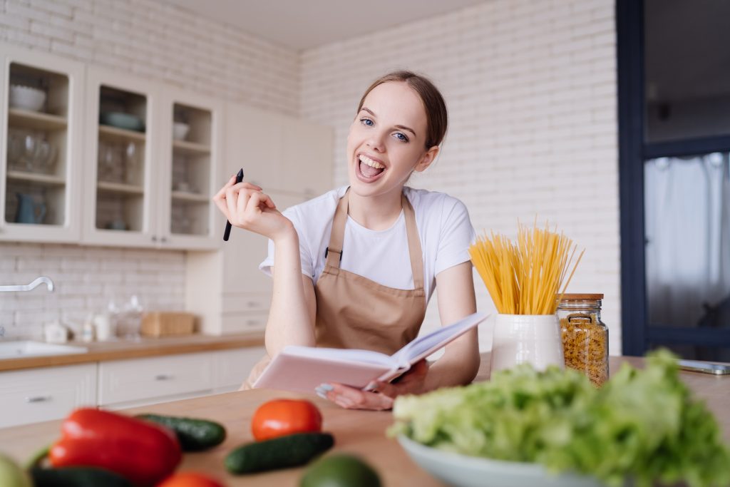 young beautiful woman kitchen apron writes down her favorite recipes fresh vegetables scaled